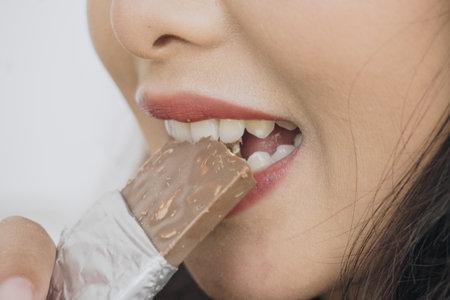 Close up portrait of Asian woman eating a chocolate bar with foil wrapper while smiling as she enjoys her snack.の写真素材
