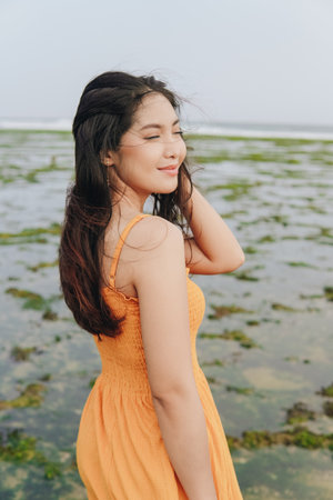 Back portrait of a young Indonesian woman wearing an orange dress enjoying the ocean view and sea...