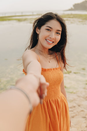 Beautiful young Asian woman wearing orange dress is smiling and holding hands with a man on a beach.の写真素材