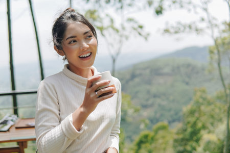 Cheerful young Asian woman holding a cup of morning coffee or tea, enjoying the view of the mountains. Nature background.の写真素材