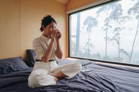Young Asian woman is sitting on a bed and drinking morning coffee or tea, enjoying her leisure time on a vacation.の写真素材