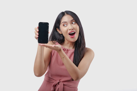 Beautiful young Asian woman wearing pink dress smiling while showing the copy space on phone screen, isolated by white background.の写真素材