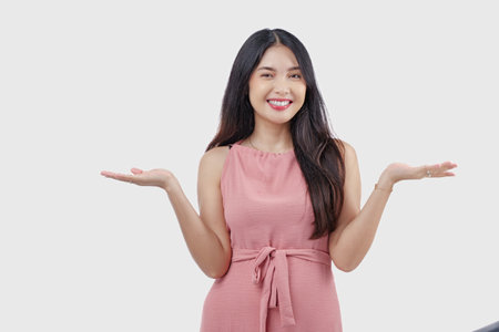 Joyful young Asian woman wearing pink dress presenting and pointing to the copy space beside her over isolated white background.の写真素材