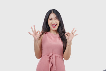 Excited young Asian woman making and showing okay sign with fingers wearing pink dress on isolated white background.の写真素材