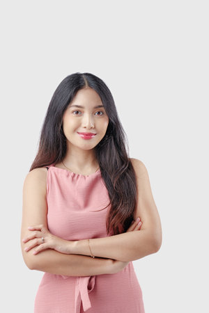 Confident young Asian woman wearing pink dress with arms crossed and happy expression posing against isolated white background.の写真素材