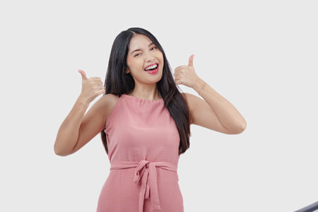 Happy Indonesian woman wearing pink dress gives thumbs up hand gesture of approval, isolated by white background.の写真素材