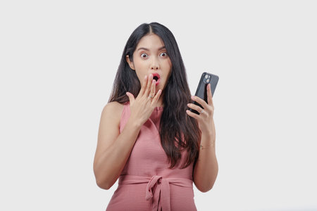 Shocked young Asian woman in pink dress holding a smartphone with a surprised expression on her...