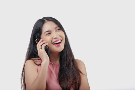 Joyful young Asian woman in a pink dress is talking on her cell phone with a happy smile over isolated white background.の写真素材