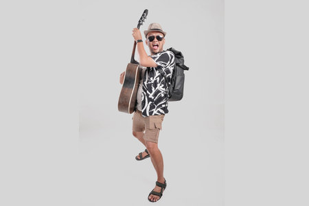 Joyful young Asian man tourist wearing backpack and sunglasses holding a guitar over isolated grey background.の写真素材