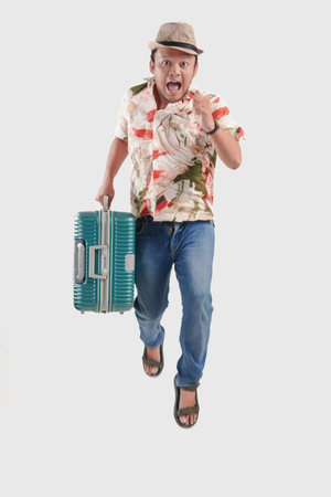 Excited young Asian man tourist wearing Hawaiian shirt jumping while bringing suitcase over isolated...