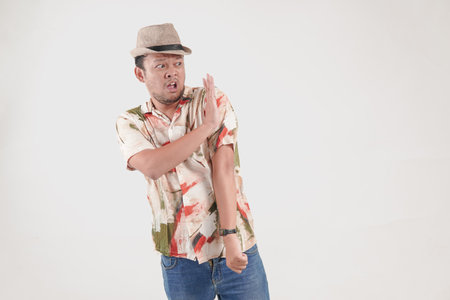 Upset Asian man wearing Hawaiian shirt and hat posing a rejection or stop hand gesture, isolated by...