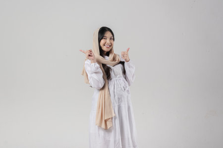 Asian Muslim pregnant woman in hijab pointing to the copy space behind her while giving thumb up gesture, isolated by white background. Ramadan concept.の写真素材