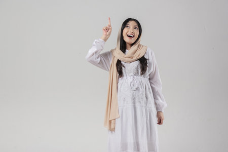 Pretty Asian Muslim pregnant woman in hijab pointing to the copy space upwards, isolated by white background. Ramadan concept.の写真素材