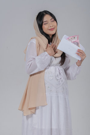 Happy mindful Asian Muslim pregnant wearing hijab smiling while holding Eid Al Fitr money gift, isolated by white background.の写真素材