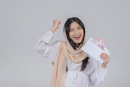 Joyful Asian Muslim pregnant woman in a veil hijab holding Eid Al Fitr money gift or THR with excited expression, isolated by white background.の写真素材