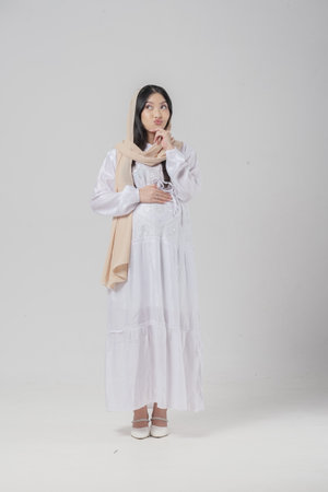 Thoughtful Asian Muslim pregnant woman wearing headscarf veil hijab focused and thinking hard, standing on isolated white background.の写真素材