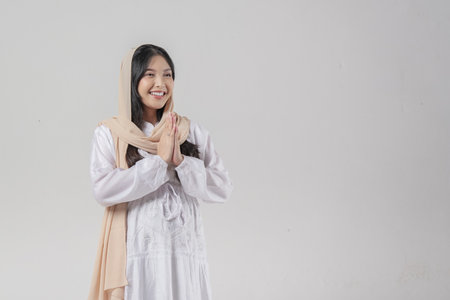 Asian Muslim woman wearing headscarf veil hijab smiling to the camera and gesturing traditional greeting isolated over white backgroundの写真素材