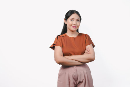 Confident young Asian woman in brown shirt and taupe pants is smiling and crossing her arms.