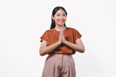 Smiling young Asian woman in brown shirt gesturing greeting or namaste isolated over white...