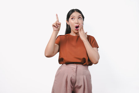 Surprised young Asian woman in a brown shirt pointing the copy space above her, isolated by white background.の写真素材