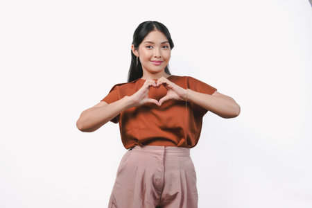 Cute young Asian woman in a brown shirt making a heart gesture using her hands, isolated over white background.の写真素材