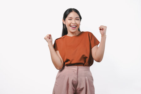 Joyful young Asian woman in a brown shirt raising her fists with happy successful expression, isolated by white background.の写真素材