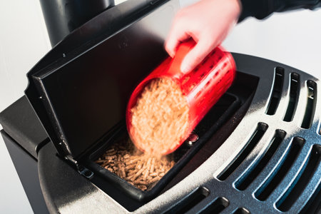 Close-up on pellets, black domestic pellet stove, man loading by hand pellets with a red 3 d printed cupの写真素材
