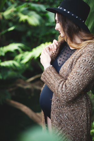Young beautiful pregnant woman in hat and cardigan among tropical plantsの写真素材