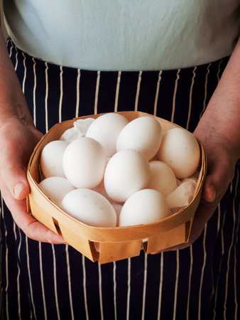 Aged woman in apron standing and holding in hands round wooden basket full of fresh raw chicken eggs, close up shot.の写真素材