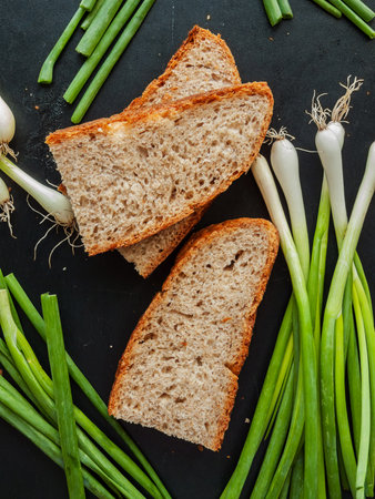 Sliced pieces of freshly baked whole wheat bread with fresh green leek onions on dark kitchen table. Traditional crusty baguette bread for healthy snack lying on table surrounded by greens.の写真素材