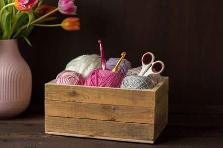 colorful woolen balls in a wooden box with crochet hooks and tulips in a vase in front of a dark brown background handmadeの写真素材