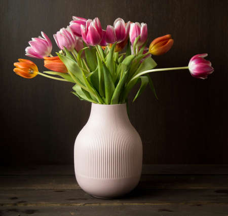 pink and orange colored tulips in vase in front of a dark brown wooden wall backgroundの写真素材