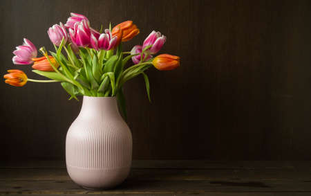 pink and orange colored tulips in vase in front of a dark brown wooden wall backgroundの写真素材