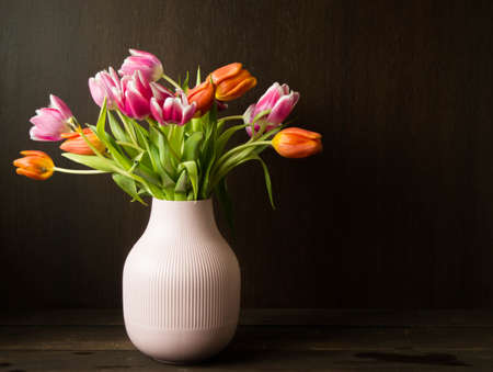 pink and orange colored tulips in vase in front of a dark brown wooden wall backgroundの写真素材