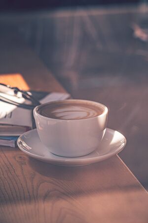 Latte coffee in white coffee cup on the wooden table in front of the large window of the coffee...