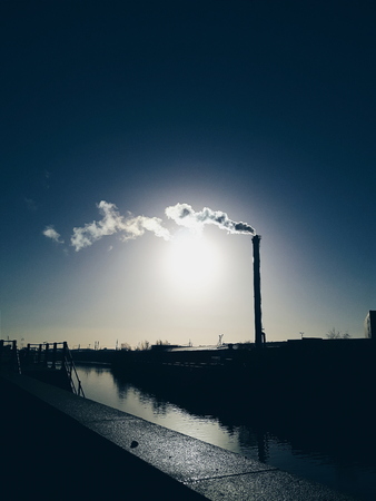 Steam rises from a smokestack on a factory silhouetted against the sunlight.の写真素材
