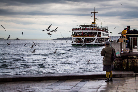 A ferry in Istanbul departing from port. Woman is standing near the water, watching and feeding the flying seagulls.のeditorial素材