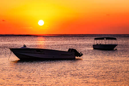 Two boats in front of the water of the setting sun are bathed in orange-red light on the island of Mauritiusの写真素材