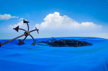 Rusty Anchor & black rope on a fishing boat covered in blue tarpaulin sheet on a beach with blue cloudy sky in the backgroundの写真素材
