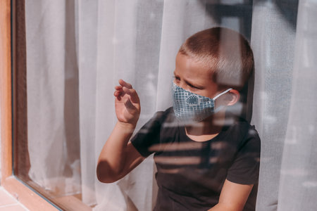 A sad little boy in a protective mask siting on the windowsill and looks out the window. A European boy draws with his fingers on the window. Stay at home quarantine   pandemic prevention.の写真素材