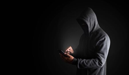 Hacker Using Smartphone. Men in black clothes with hidden face looks at smartphone screen on black background with copy space. with clipping path.の写真素材