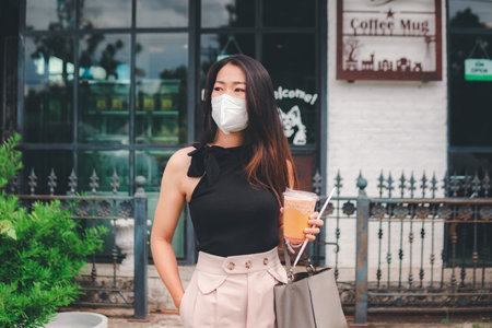 A beautiful asian woman holding glass of coffee open the door walk out cafe wear white maskの写真素材