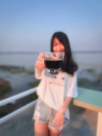 Beautiful Asian woman standing and drinking coffee by the river in the morning.の写真素材