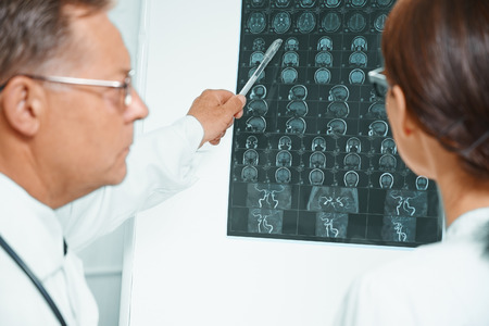 Senior doctor examines MRI image of human head. Older man doctor teaches young woman doctorの写真素材