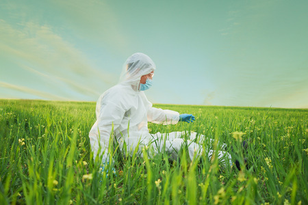 Scientist man in biohazard uniform sits on green summer meadow and touches green grassの写真素材