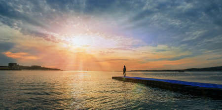 Woman standing on pier near the sea and looking at sunset, tranquil scene. Beautiful seascape. Image with sunlight effectの写真素材