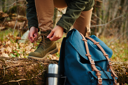 Female hiker tying shoelaces outdoors in autumn forest, near thermos and backpack. View of legs. Hiking and leisure themeの写真素材