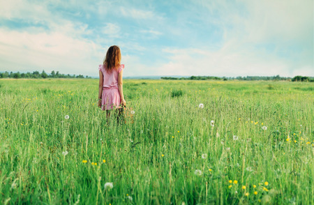 Little girl in pink dress standing with suitcase on summer green meadow, rear viewの写真素材