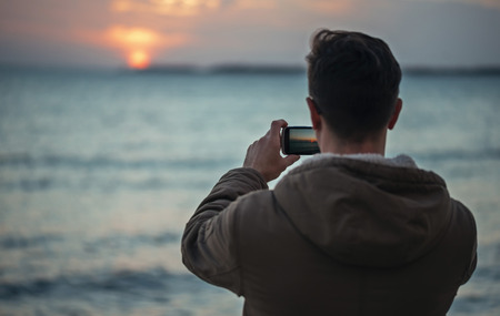 Traveler young man takes photographs beautiful sunset over the sea, rear viewの写真素材