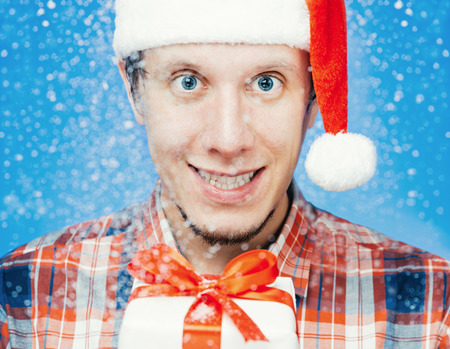 Portrait of cheerful young man in santa hat with a gift box under snow. New Year or Christmas themeの写真素材